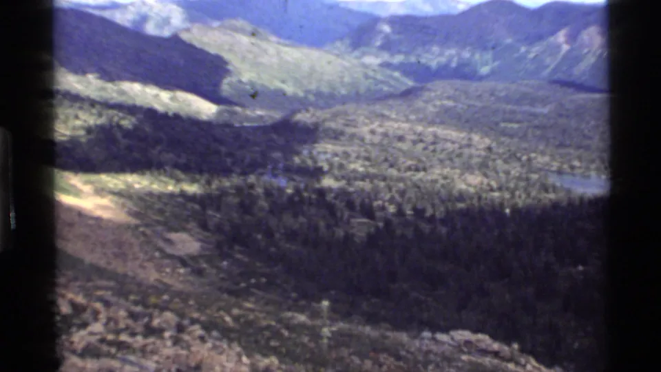 1972 8mm landscape near Holland Lake, Montana, with rocky foreground, evergreen forest, open slopes, and distant water.