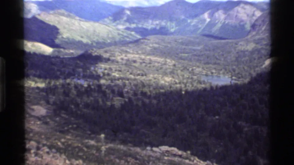 Handheld 1972 Holland Lake overlook frame with forested slopes, a blue lake, and layered Montana mountains.