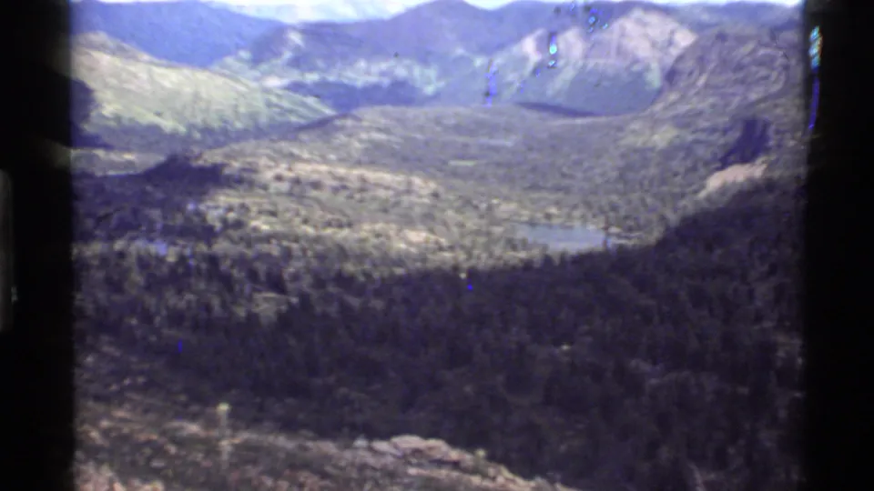 1972 home-movie frame looking over Holland Lake and a tree-lined Montana valley from a high rocky viewpoint.