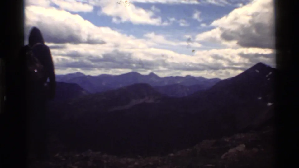 1972 8mm frame from Holland Lake, Montana, with a backpacked figure silhouetted against dark mountain ridges and cloud cover.