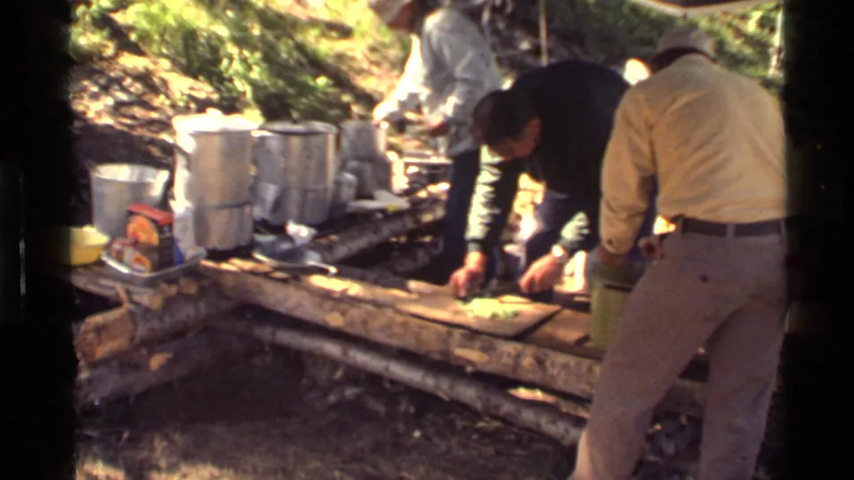 Men in casual 1970s clothing work at a rustic outdoor camp setup with aluminum cookware and logs in Montana.