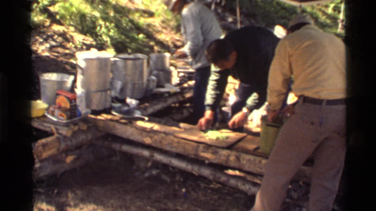 Men preparing food at a rustic outdoor camp counter with pots and gear nearby.