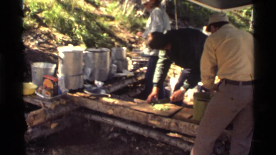 At the Montana campsite, men handle food at a rough wooden counter with large aluminum pots and a tarp shelter visible.