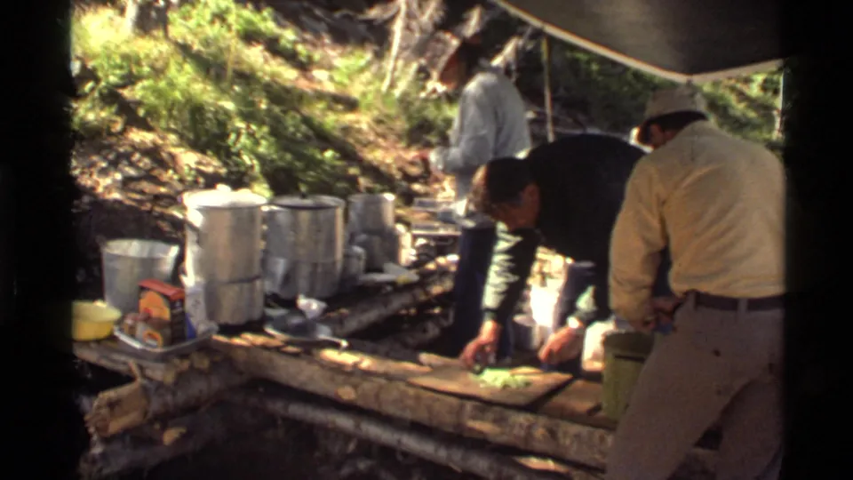 Men at Holland Lake prepare food beneath a tarp, surrounded by cookware, a wooden counter, and sunlit forest vegetation.