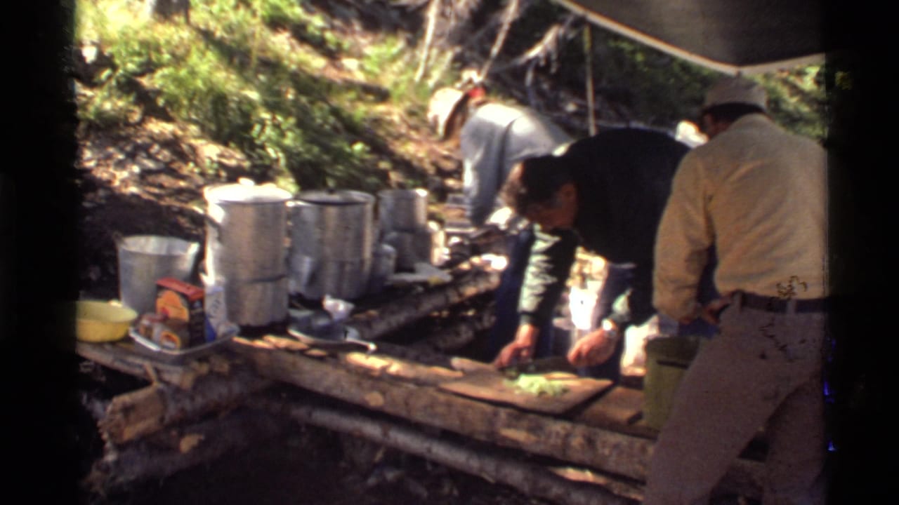 1972 Holland Lake, Montana home-movie frame showing men preparing food under a tarp at a rustic campsite.