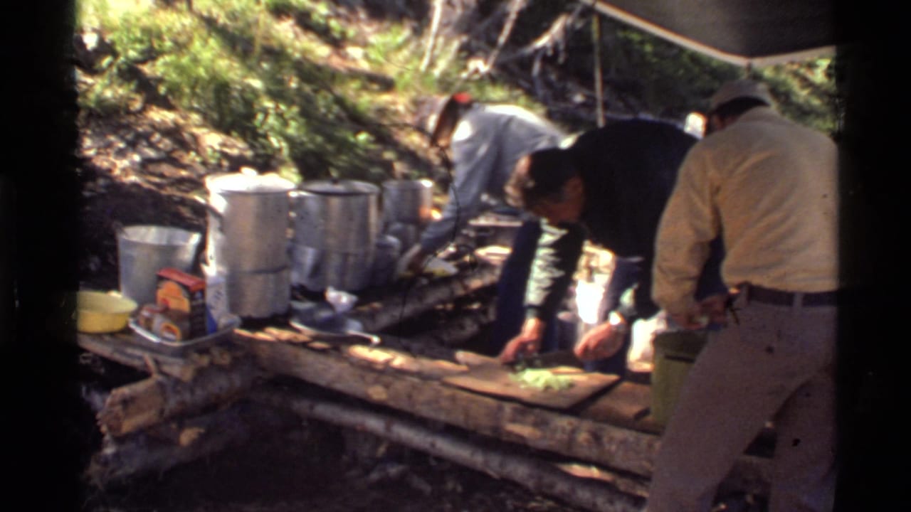 Men at a 1972 Montana forest campsite work around a log table with cookware, vegetables, and packaged supplies.