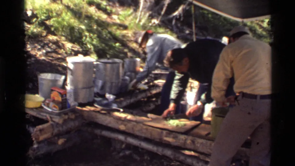 Men at a 1972 Montana forest campsite work around a log table with cookware, vegetables, and packaged supplies.