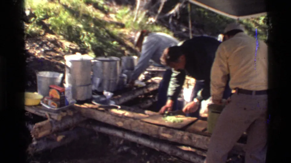 1972 Holland Lake camping scene with men preparing vegetables at a rough table under filtered forest light.