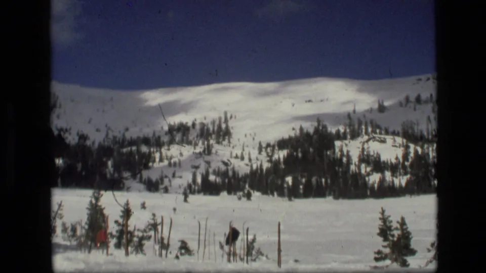 1972 Idaho 8mm frame showing a broad snowy slope with evergreens and small distant ski-area details near the lower left.