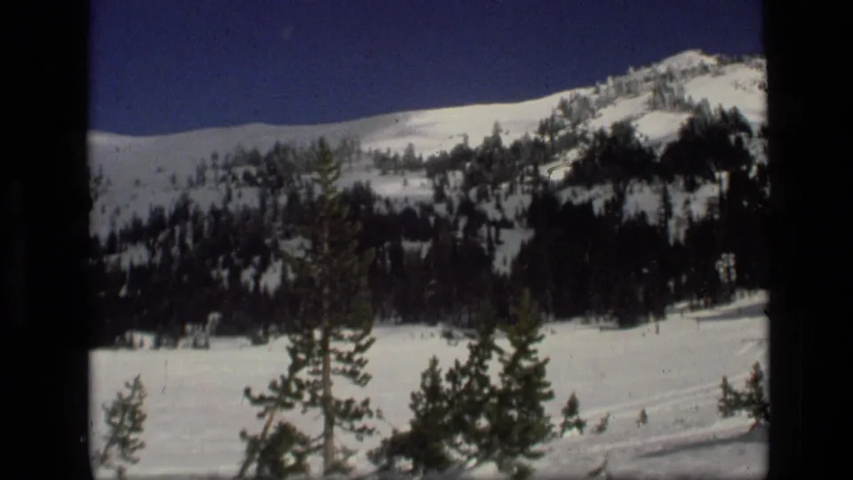 1972 Idaho winter scene with a snow-covered hillside, scattered evergreens and a small distant structure or object at the base.