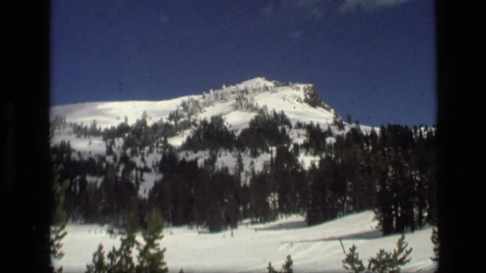 1972 Idaho home-movie frame of a snowbound mountain slope, evergreen forest, and blue sky with small clouds.