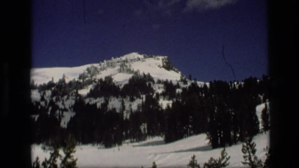 1972 Idaho 8mm frame with dark evergreens crossing a snow-covered slope below a blue sky.