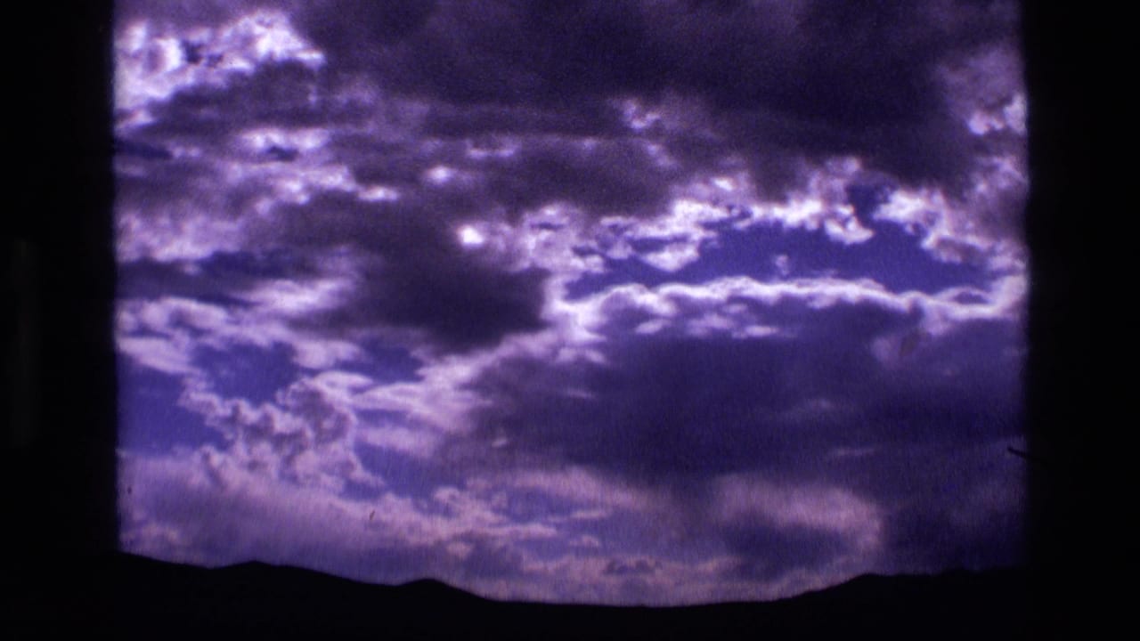 1972 Idaho 8mm frame showing dense purple-blue clouds with bright openings above a silhouetted mountain horizon.
