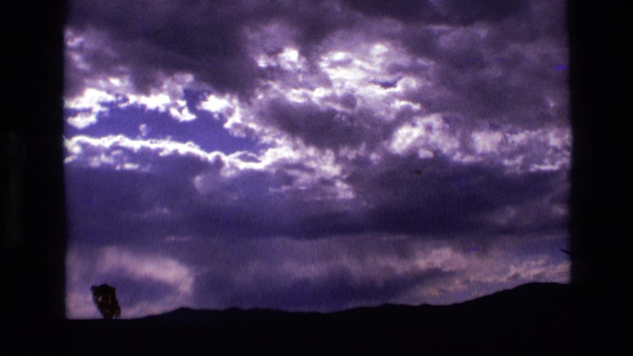 1972 Idaho 8mm frame with turbulent purple clouds, a dark horizon, and a small indistinct silhouette near the lower center.
