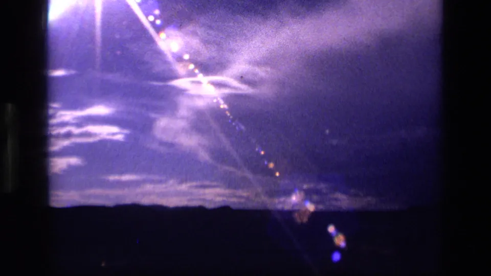 1972 Idaho 8mm frame with diagonal flare streaks, wispy clouds, and a dark mountain line at the bottom.