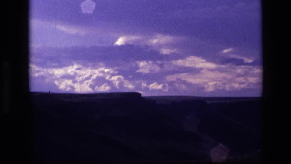 1972 Idaho 8mm frame with layered stormlike clouds, a dark ridge silhouette, and lens flare near the lower edge.