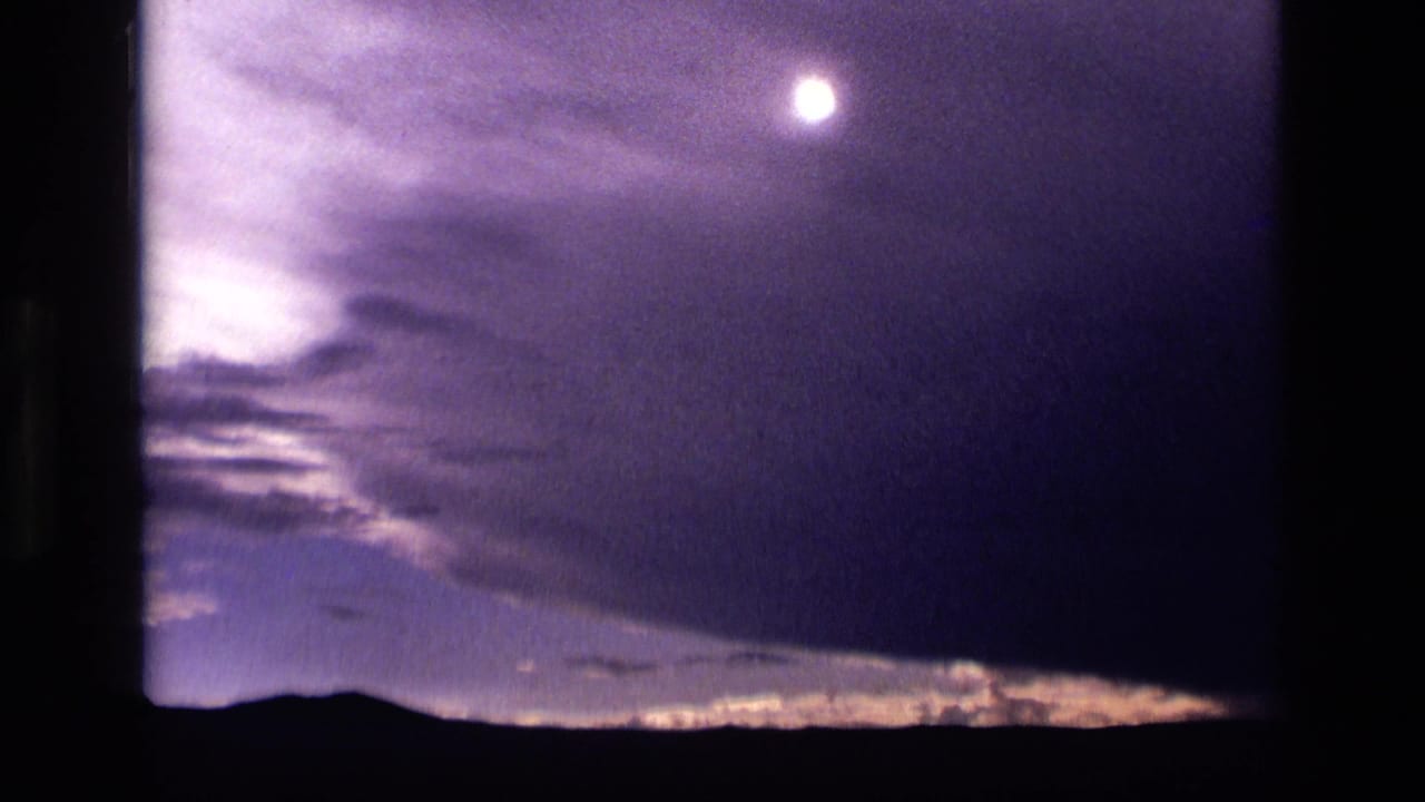 1972 Idaho 8mm frame showing a bright moon, layered dark clouds, and a low horizon under a blue-purple night sky.