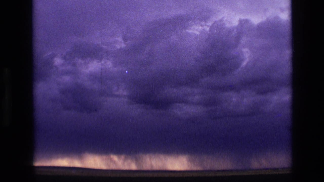 1972 Idaho 8mm frame with a broad, dark cloud mass and faint vertical streaks near the horizon that may be distant rain.