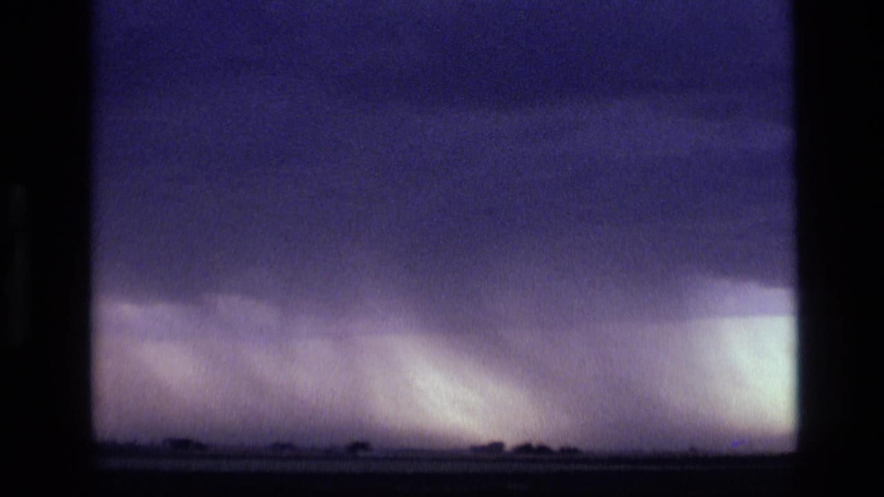 1972 Idaho 8mm frame showing dark purple storm clouds above a low horizon and bright rain shaft in the distance.