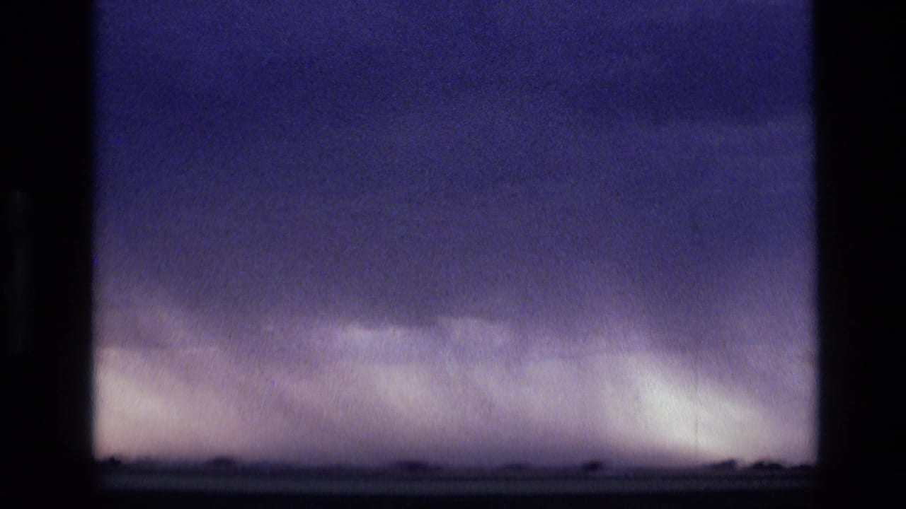 1972 Idaho home-movie frame with dense storm clouds, pale rain near the horizon, and sparse dark landforms below.