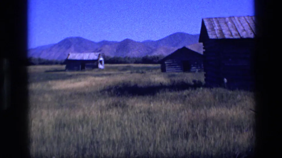 1972 Idaho prairie view with rustic log cabins, open grass, and distant mountains under a blue sky.