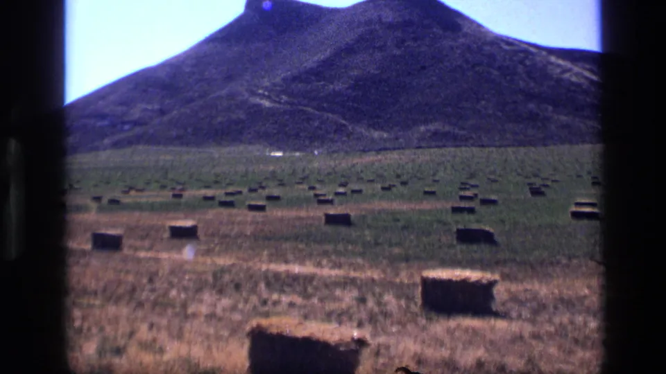 1972 Idaho home-movie frame of square hay bales across a broad field, with a dark hillside and pale blue sky behind them.