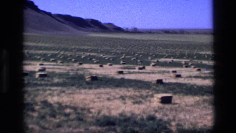 1972 Idaho 8mm frame of a broad hay field with scattered rectangular bales, distant low hills, and a dark film edge.