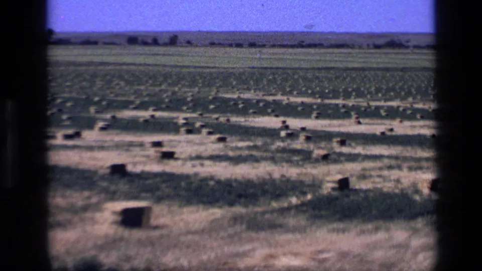 1972 Idaho home-movie frame showing a dry field with hay bales, green agricultural rows, and a distant tree line under blue sky.