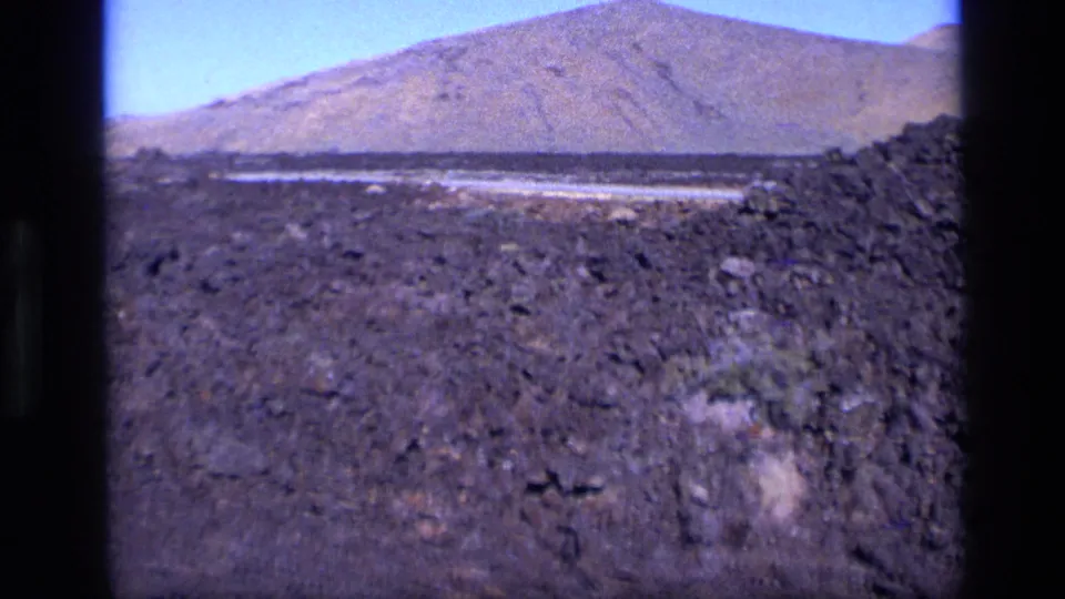 1972 Idaho home-movie frame showing rocky ground leading toward a low mountain ridge beneath a clear pale sky.