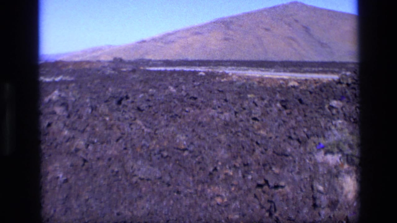 Dark lava rock field with distant mountain under blue sky.