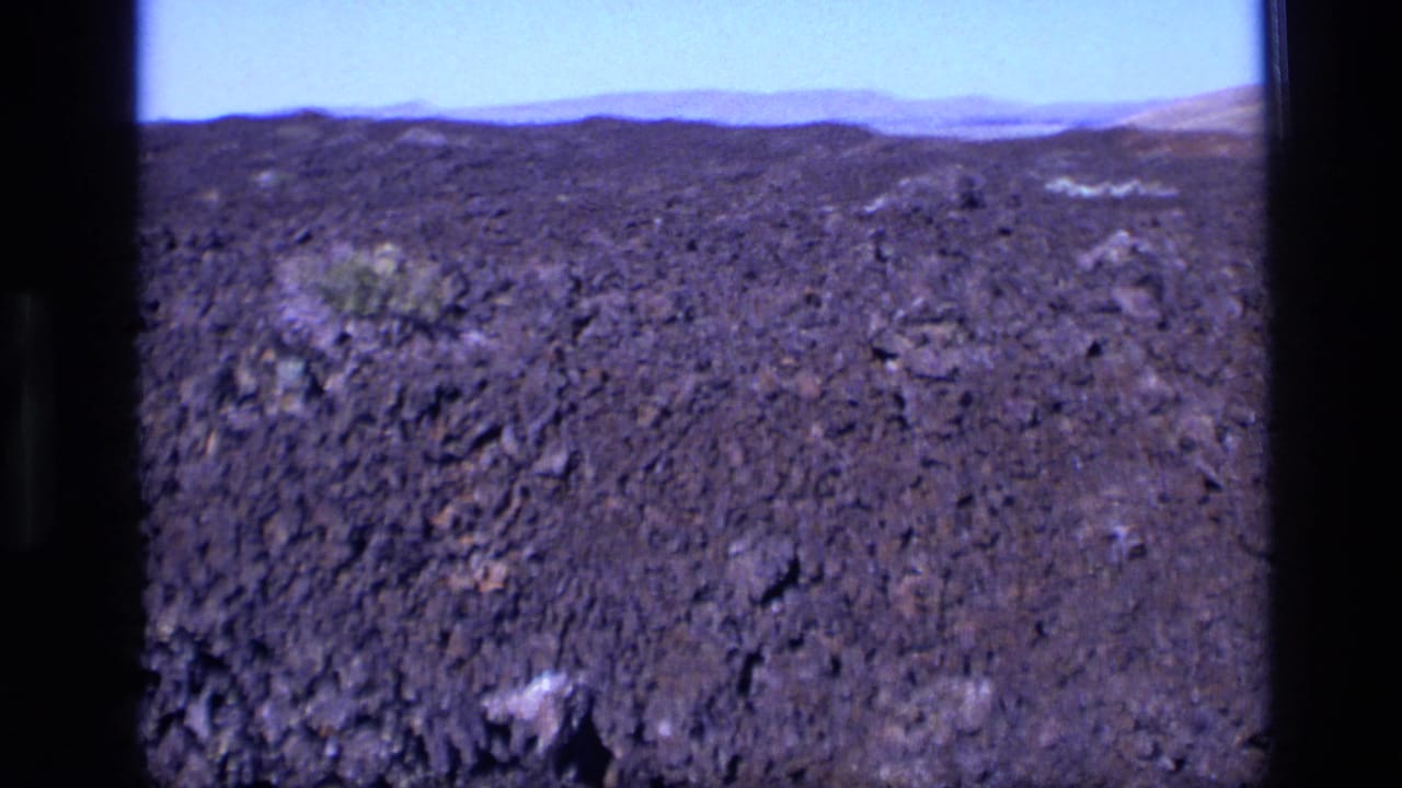1972 Idaho 8mm frame showing dark porous volcanic terrain, low hills, pale sky, and scattered small shrubs.