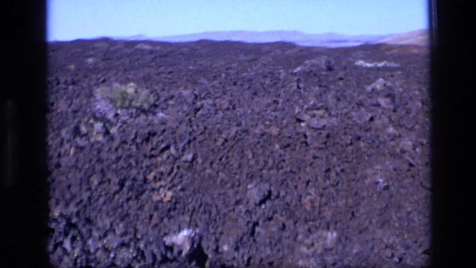 1972 Idaho 8mm frame showing dark porous volcanic terrain, low hills, pale sky, and scattered small shrubs.