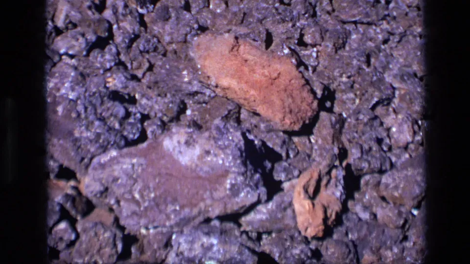 Close 1972 Idaho 8mm view of a reddish-brown mineral-like stone resting amid darker porous rocks.