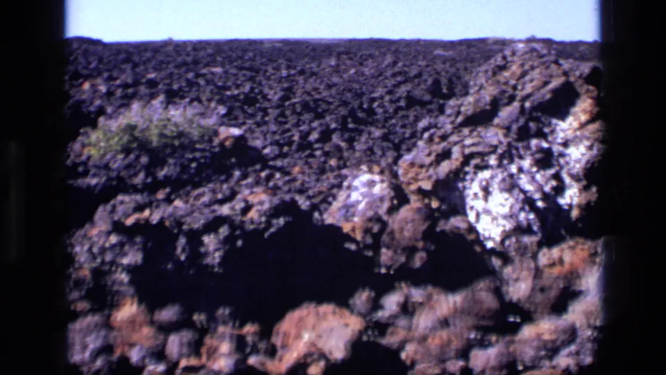 1972 Idaho 8mm frame showing rugged dark rocks, a scrub plant, and lighter mineral deposits in bright daylight.