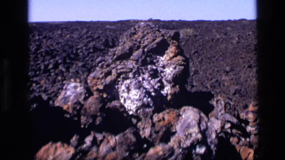 1972 Idaho 8mm frame of a large dark rock formation with pale mineral deposits set against rocky ground and sky.