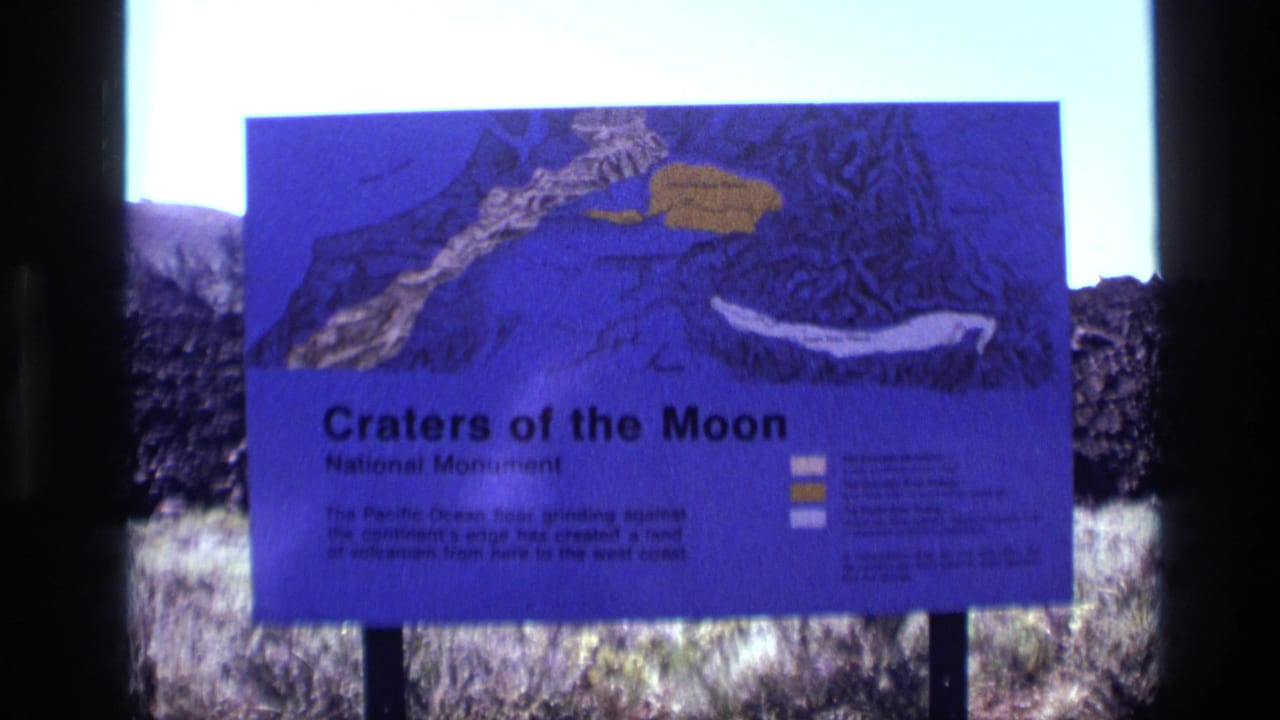 Purple Craters of the Moon sign beside dark volcanic rock and dry grass.