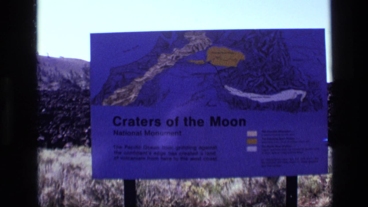 Visitor-shot 8mm view of the Craters of the Moon National Monument sign in Idaho, with volcanic rock and sparse vegetation behind it.