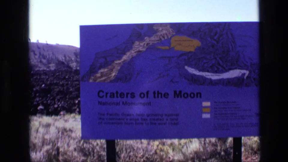 1972 home-movie frame of the Craters of the Moon sign, showing its illustrated map and text against the volcanic Idaho setting.