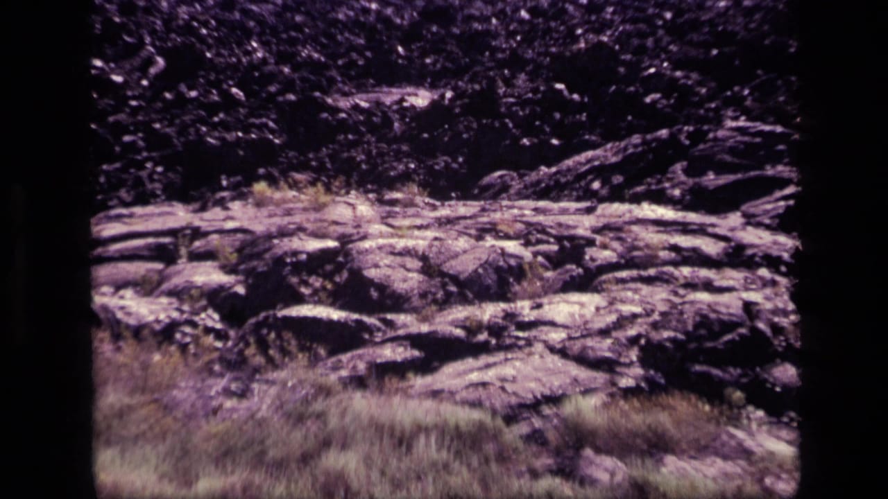 1972 Idaho 8mm frame showing rocky, fire-scorched ground with dry grass and dark vegetation in the background.