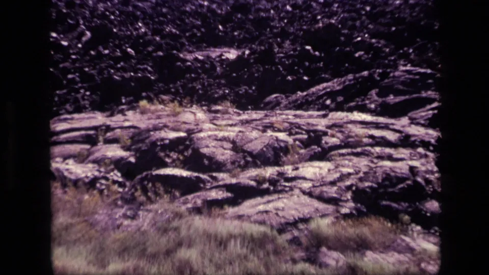 1972 Idaho 8mm frame showing rocky, fire-scorched ground with dry grass and dark vegetation in the background.