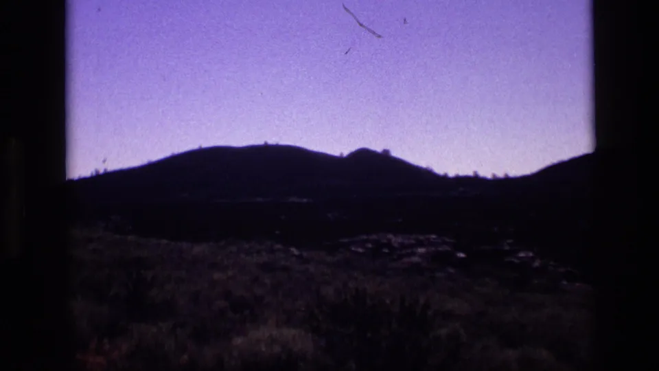 1972 Idaho 8mm frame with a dark scorched field, dry grasses, and low hills under a purple-tinted sky.