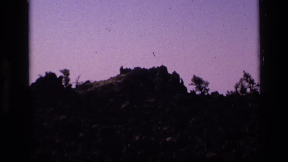 1972 Idaho home-movie frame showing a jagged rocky outcrop with sparse trees under a purple sky.