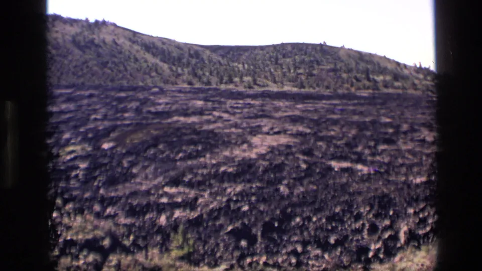 1972 Idaho home-movie frame with scorched-looking or volcanic-looking ground before a wooded hillside.