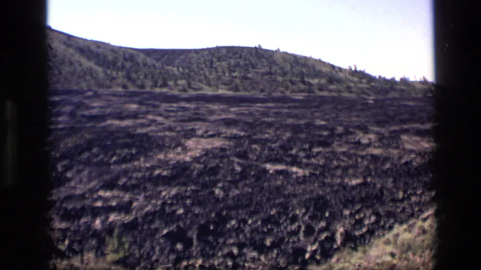 1972 Idaho 8mm frame showing rough dark ground, scattered vegetation, and a wooded ridge near the end of the panoramic move.