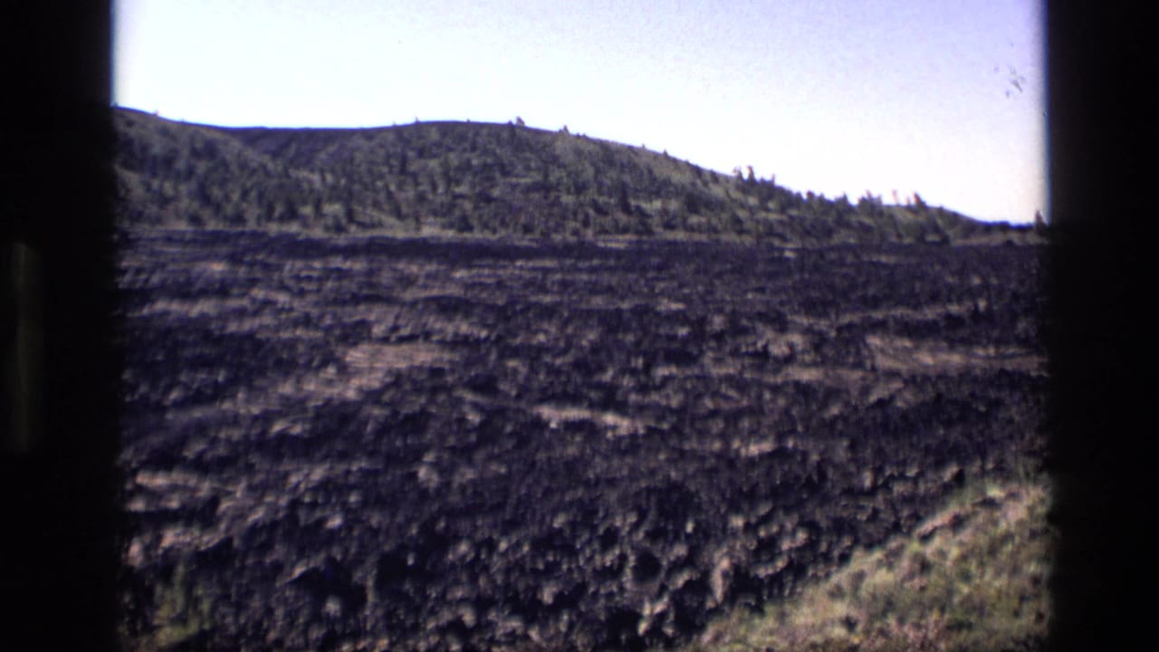 1972 Idaho 8mm frame showing dark textured ground, distant low hills, sparse trees, and a pale sky.