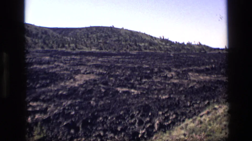 1972 Idaho 8mm frame showing dark textured ground, distant low hills, sparse trees, and a pale sky.