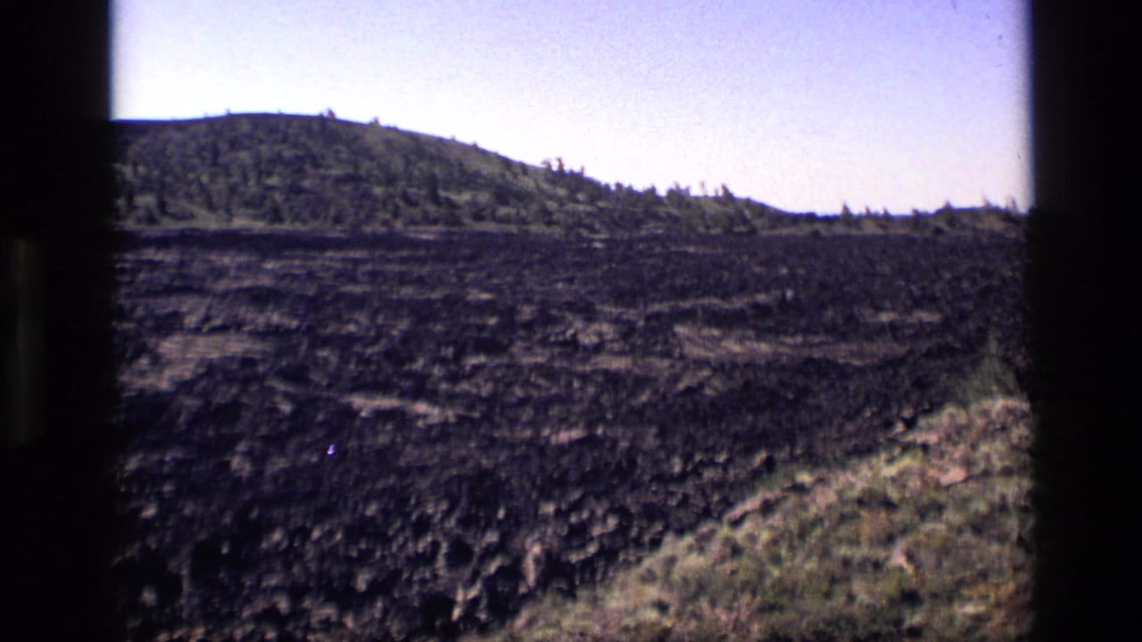 1972 Idaho home-movie frame with dark rough terrain, a low wooded ridge, and visible 8mm gate borders.