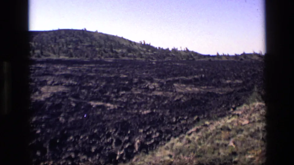 1972 Idaho home-movie frame with dark rough terrain, a low wooded ridge, and visible 8mm gate borders.