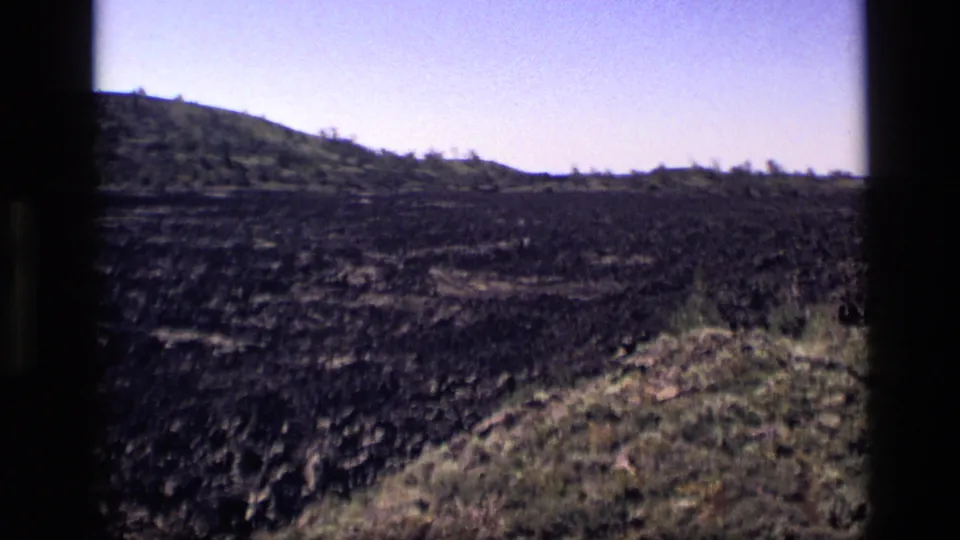 1972 Idaho 8mm view of dark uneven ground, grassy roadside edge, distant low hills, and open sky.