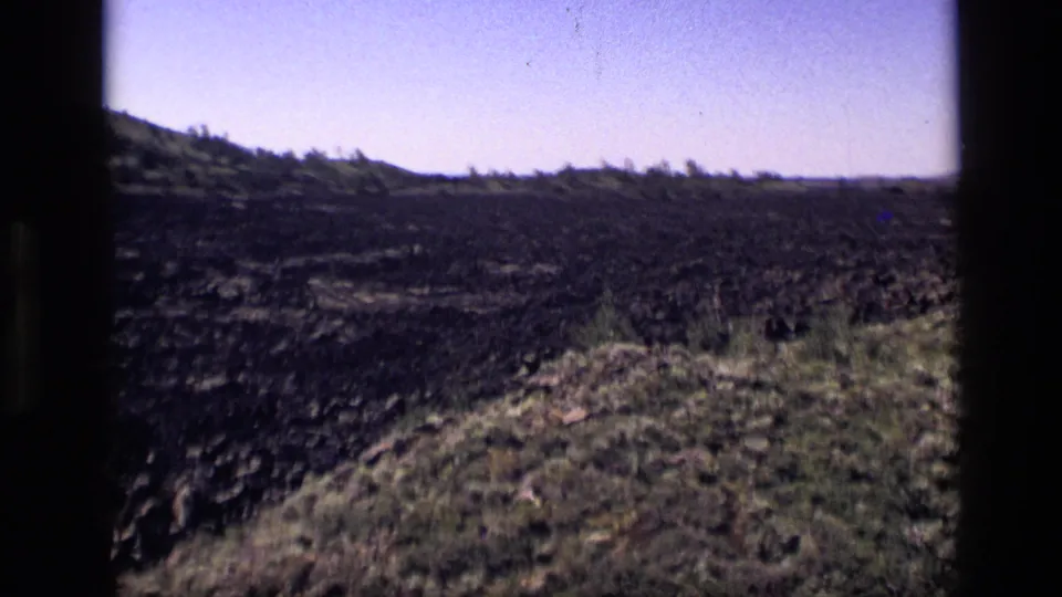 1972 Idaho frame showing a grassy slope beside dark rocky terrain, with low hills and pale sky beyond.